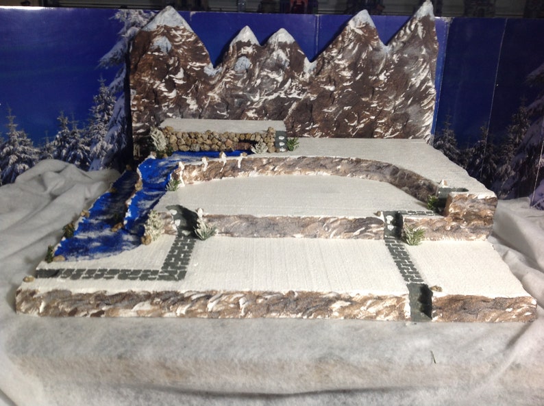 May include: A miniature winter scene with a blue river, snow-covered ground, and a backdrop of brown mountains. The scene is built on a multi-level platform with stone walls and paths.