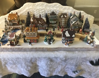 May include: A miniature Christmas village scene with a snowy mountain backdrop, a river, and several houses with lit windows. The village includes a church, a bakery, a pub, and a general store. There are also several small trees and figures of people walking around the village.