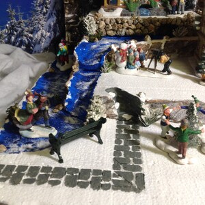 May include: A miniature winter village scene with a blue painted stream, a small park bench, and several figurines of people and snowmen. The scene is set on a white surface with a gray stone path.