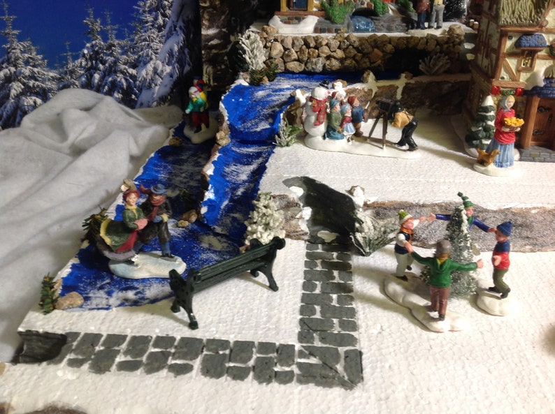 May include: A miniature winter village scene with a blue painted stream, a small park bench, and several figurines of people and snowmen. The scene is set on a white surface with a gray stone path.
