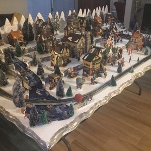 May include: A miniature Christmas village scene with white snow-covered mountains, a winding river, and a variety of houses and buildings. The village is lit with small lights and includes miniature trees and figures.
