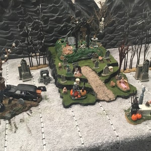 May include: A Halloween village scene with a graveyard, a spooky tree, a hearse, and a skeletal figure. The scene is set on a white surface with a black background.