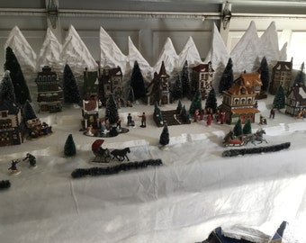 May include: A miniature Christmas village scene with white snow-capped mountains, houses, trees, and figures. The village is set on a white tablecloth with a black garland.