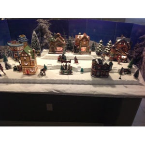 May include: A miniature Christmas village scene with snow-covered houses, trees, and figures. The houses are decorated with lights and have a festive feel. The scene is set on a white base with a dark brown background.