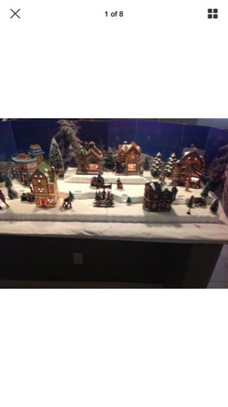 May include: A miniature Christmas village scene with snow-covered houses, trees, and figures. The houses are decorated with lights and have a festive feel. The scene is set on a white base with a dark brown background.