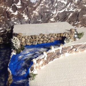 May include: A miniature winter scene with a small stone bridge over a blue river. The river is surrounded by white snow and there are small trees on either side of the bridge.