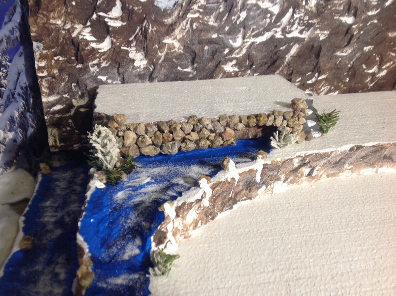 May include: A miniature winter scene with a small stone bridge over a blue river. The river is surrounded by white snow and there are small trees on either side of the bridge.