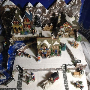 May include: A miniature Christmas village scene with snow-covered houses, a river, and figures. The village is set against a backdrop of mountains and a starry night sky.