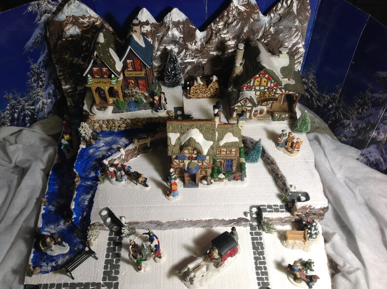 May include: A miniature Christmas village scene with snow-covered houses, a river, and figures. The village is set against a backdrop of mountains and a starry night sky.