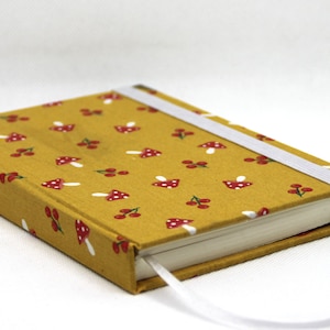 Könnte beinhalten: Ein rechteckiges Notizbuch mit einem senfgelben Stoffeinband, der mit einem sich wiederholenden Muster aus kleinen roten Pilzen und Kirschen bedruckt ist. Das Notizbuch wird mit einem weißen Gummiband verschlossen und hat ein weißes Lesebändchen.