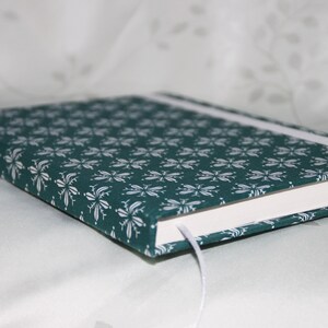 May include: A teal blue notebook with a white floral pattern on the cover. The notebook has a white ribbon bookmark and a white spine.