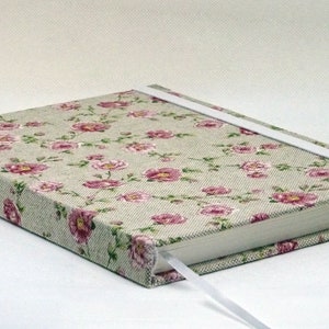 May include: A light grey journal with a floral pattern and a white ribbon closure. The cover features a repeating pattern of pink flowers and green leaves.