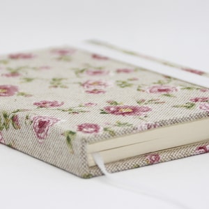 Könnte beinhalten: Ein rechteckiges Notizbuch mit einem beigen Stoffeinband, der mit einem sich wiederholenden Muster aus rosa Blumen und grünen Blättern bedruckt ist. Das Notizbuch hat ein weißes Gummiband als Verschluss und ein schmales weißes Lesebändchen.