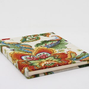 Puede incluir: Un diario cerrado con una cubierta de diseño floral. La cubierta presenta un fondo blanco con patrones florales rojos, naranjas, amarillos, verdes y azules.