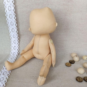 May include: A beige fabric doll with a button on its arm, sitting on a beige fabric surface. The doll has a simple design and is unfinished.