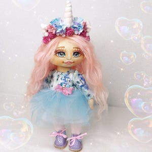Puede incluir: Una muñeca hecha a mano con cabello rosado, un vestido floral azul y blanco, una falda de tul azul y una diadema de cuerno de unicornio con flores rosas y azules.