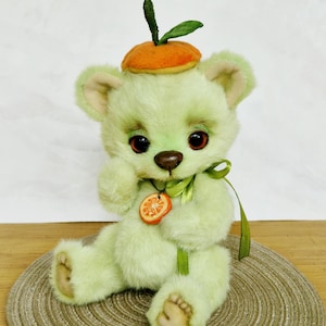 Può includere: Un orsacchiotto fatto a mano, verde chiaro, con un cappello arancione stravagante e un ciondolo a forma di fetta d'arancia abbinato. L'orso ha occhi rosso scuro, un naso marrone ed è seduto su un tappetino intrecciato. Un nastro verde è legato intorno al collo.