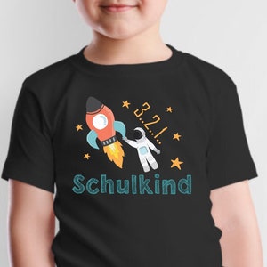 Könnte beinhalten: Schwarzes T-Shirt mit einem Aufdruck eines Astronauten in einer Rakete mit Sternen und dem Text "3...2...1! Schulkind" in Blau.