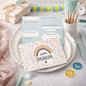 May include: A rectangular card with a white background featuring a colorful confetti pattern and a central rainbow graphic above the text 'meine EINSCHULUNG'. Behind it, a light blue questionnaire card with German text and blank lines for writing.