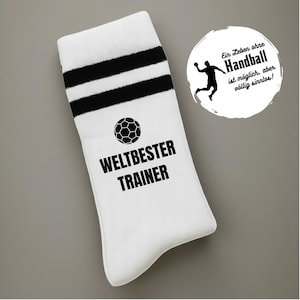 May include: White athletic sock with two black stripes at the top and a soccer ball graphic. The words "WELTBESTER TRAINER" are printed in black. A circular graphic with a handball player and German text is also present.