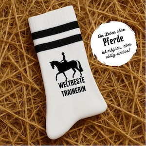 Puede incluir: Calcetines deportivos blancos con rayas negras y un gráfico de equitación. Los calcetines presentan una silueta de caballo y jinete, con las palabras "WELTBESTE TRAINERIN" debajo. Una burbuja de texto circular dice "Ein Leben ohne Pferde ist möglich, aber völlig sinnlos!"