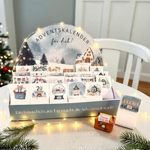 Op de afbeelding: Een adventskalender met een winterse scène. De kalender heeft kleine deuren met nummers 1-24, elk met een unieke illustratie. De tekst "ADVENTSKALENDER für dich!" staat bovenaan. Een kleine chocoladetraktatie ligt ernaast.