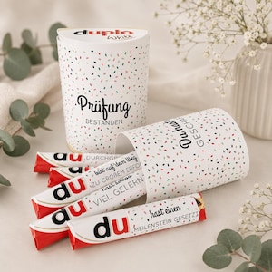 Pode incluir: Uma caixa de presente branca e um recipiente cilíndrico com confetes coloridos. A caixa diz "Prüfung Bestanden" e o recipiente "Du hast Geschafft". Várias barras de chocolate com a palavra "du" são visíveis.