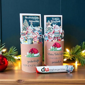 Könnte beinhalten: Zwei weihnachtliche Papierarbeiten mit Weihnachtsmann und Kindern. Die Arbeiten tragen die Texte "Wir wünschen frohe Weihnachten" und "Ein kleiner Weihnachtsgruß". Eine Süßwarenriegel mit dem Text "du bringst uns zum lachen" liegt davor.