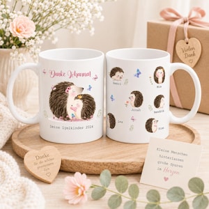 Op de afbeelding: Twee witte keramische mokken met egel illustraties. E&eacute;n mok heeft een ouder en kind egel met de tekst "Danke Johanna!" en "Deine Igelkinder 2024." De andere mok heeft individuele egel ontwerpen met namen. Een houten hart en een geschenkdoos zijn ook zichtbaar.