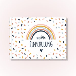 May include: A rectangular book cover with a white background featuring a pattern of small, scattered confetti shapes in shades of pink, blue, and yellow. Centered is a rainbow graphic above the text "meine EINSCHULUNG" and a small heart.