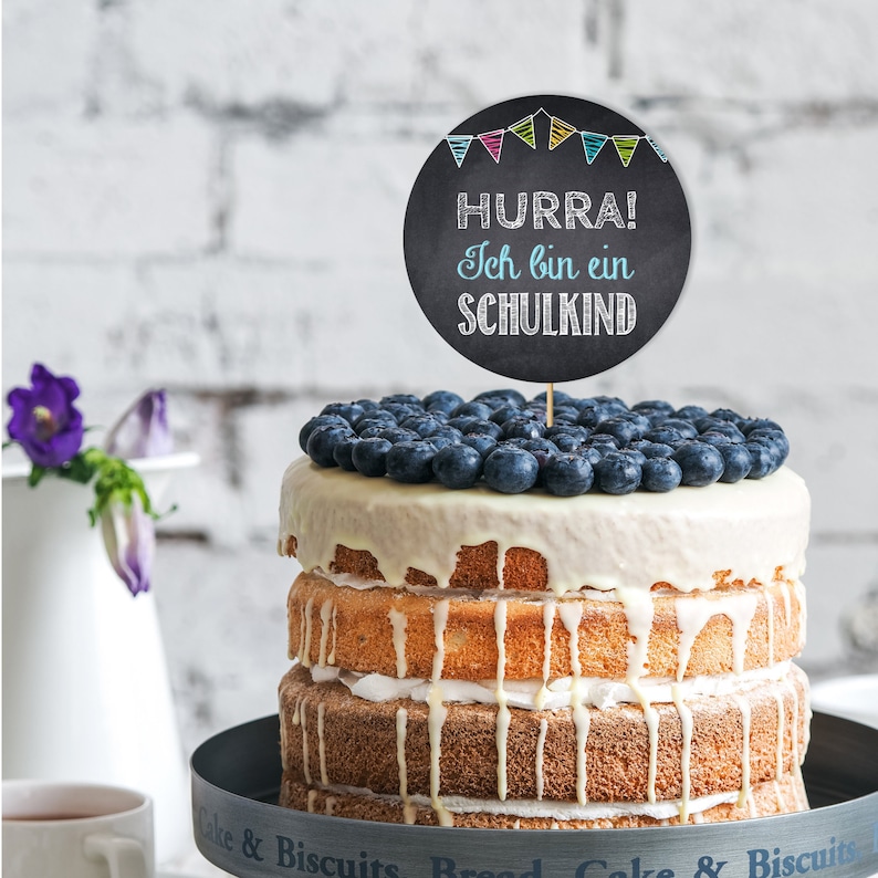 Peut inclure: G&acirc;teau &agrave; trois &eacute;tages avec gla&ccedil;age blanc et myrtilles. Un cake topper rond en tableau noir indique "HURRA! Ich bin ein SCHULKIND" avec des fanions color&eacute;s. Le g&acirc;teau est pos&eacute; sur un pr&eacute;sentoir &agrave; g&acirc;teau gris fonc&eacute;.