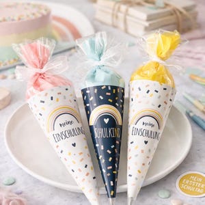 May include: Three cone-shaped gift bags with clear plastic tops tied with white ribbons. The bags feature a confetti pattern, a rainbow graphic, and the German text "meine EINSCHULUNG" or "SCHULKIND". The bags are pink, light blue, and yellow.