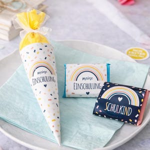 May include: A cone-shaped gift bag and two rectangular chocolate bars featuring a rainbow graphic and the German text "meine EINSCHULUNG" and "SCHULKIND". The items are decorated with colorful confetti patterns on white and dark blue backgrounds.