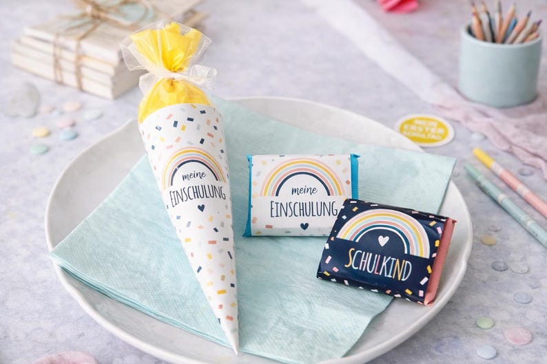 May include: A cone-shaped gift bag and two rectangular chocolate bars featuring a rainbow graphic and the German text "meine EINSCHULUNG" and "SCHULKIND". The items are decorated with colorful confetti patterns on white and dark blue backgrounds.