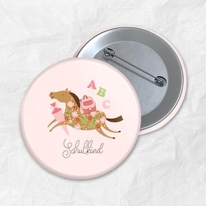 Puede incluir: Botón rosa con un caballo de dibujos animados que lleva una mochila escolar y un cono. El caballo está decorado con flores. El texto "Schulkind" está escrito debajo del caballo.