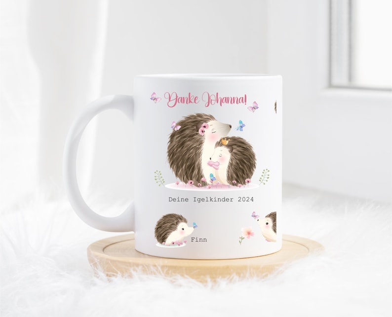 Op de afbeelding: Witte keramische mok met een illustratie van twee egels, vlinders en bloemen. De mok heeft de tekst "Danke Johanna!" en "Deine Igelkinder 2024". De naam "Finn" is ook afgebeeld op de mok.