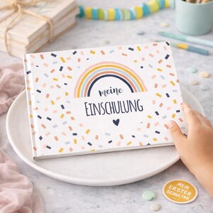 May include: A rectangular book with a white cover featuring a colorful rainbow graphic, scattered confetti-like shapes in orange, blue, and yellow, and the text "meine EINSCHULUNG" in dark blue lettering with a small heart icon below the text.