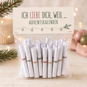 Op de afbeelding: Een doorzichtig plastic zakje met talloze witte papierrollen die zijn vastgezet met kleine gekleurde elastiekjes. De bovenkant van het zakje heeft een kartonnen label met de tekst "ICH LIEBE DICH, WEIL ... ADVENTSKALENDER" en een illustratie van hartjes.