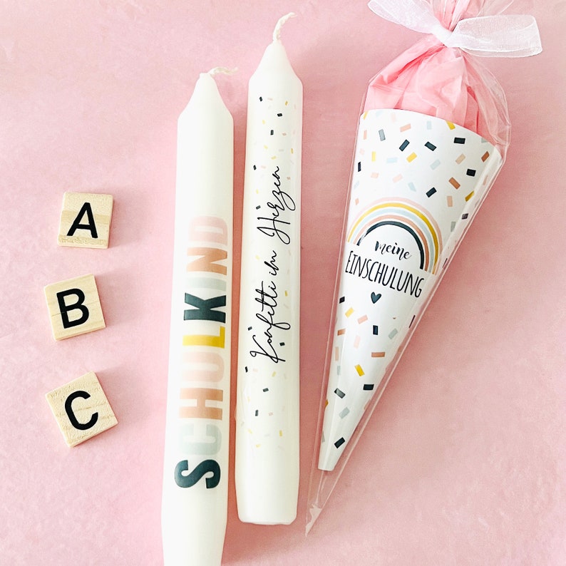 May include: Two white taper candles and a cone-shaped gift bag. One candle features the text SCHULKIND in pastel colors, the other has Konfetti im Herzen written in black. The cone bag displays a rainbow and the text meine EINSCHULUNG.