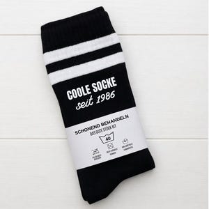Peut inclure: Chaussettes rayées noires et blanches avec le texte "COOLE SOCKE seit 1986". Les chaussettes ont une bande blanche en haut et une étiquette blanche avec des instructions de lavage.