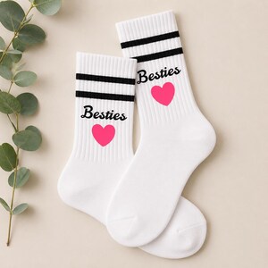 Puede incluir: Un par de calcetines deportivos blancos con rayas negras y la palabra "Besties" en escritura negra, junto con un coraz&oacute;n rosa. Los calcetines est&aacute;n hechos de un material suave.