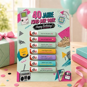 Puede incluir: Una tarjeta de cumpleaños de 40 años con gráficos nostálgicos de los 90, incluyendo una consola portátil, un Tamagotchi, una zapatilla de plataforma, un Discman y un radiocasete. Siete barritas de chocolate llevan textos en alemán.