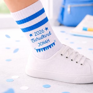 Op de afbeelding: Witte sportsokken met blauwe strepen en de tekst "2026 Schulkind JONAH" in blauw. De sokken worden gedragen met witte sneakers. Blauwe confetti ligt verspreid rond de schoenen.