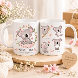 May include: Two white ceramic mugs with koala bear illustrations and floral accents. One mug features a mother koala with a baby, the other has multiple koala designs. Text includes names and a message.