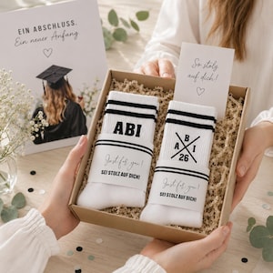 Könnte beinhalten: Geschenkbox mit zwei Paar weißen Socken mit schwarzen Streifen und dem Aufdruck "ABI". Die Socken haben ein Band mit der Aufschrift "Just for you! SEI STOLZ AUF DICH!". Eine Karte und eine Abschlussankündigung befinden sich ebenfalls in der Box.
