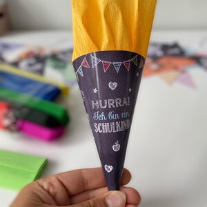 May include: A purple paper cone with the text "HURRA! Ich bin ein SCHULKIND" and decorative drawings, filled with yellow tissue paper. The cone has a festive design with colorful flags, stars, hearts, and an apple. The cone is held by a hand.