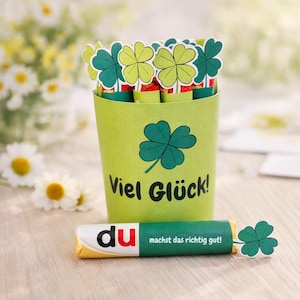 Può includere: Un contenitore verde chiaro con un design a quadrifoglio e la scritta "Viel Glück!" contiene diverse barrette di cioccolato sormontate da ritagli di trifogli. Una singola barretta di cioccolato con la parola "du" e la frase "machst das richtig gut!" è davanti.