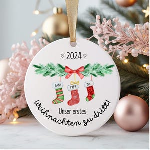 Könnte beinhalten: Runder weißer Weihnachtsbaumschmuck mit einer Aquarell-Illustration von drei Strümpfen, die an einer grünen Girlande hängen. Die Strümpfe sind mit "Mama", "Papa" und "Mia" beschriftet. Der Anhänger enthält außerdem den Text "2024", "Unser erstes Weihnachten zu dritt!" und eine rote Schleife.