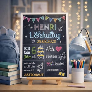 Può includere: Una lavagna con il nome "HENRI" e il testo "1. Schultag" in scrittura bianca. Il cartello include disegni di una torta, un razzo e un cuore, con la data 29.08.2020.