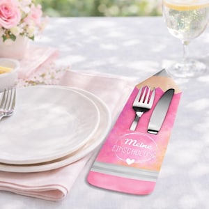 May include: A pencil-shaped cutlery holder with a fork and knife. The holder is pink with a watercolor effect and the text "Meine Einschulung" in a white circle. The setting includes white plates, a glass of water, and a floral arrangement.